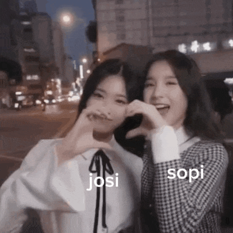 two girls are making a heart shape with their hands while standing next to each other on a city street .