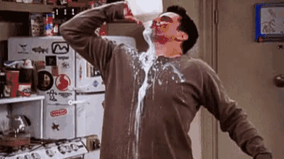 a man is drinking milk from a container in a kitchen with milk pouring out of his shirt .