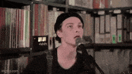 a young man in a black hat is singing into a microphone in front of a bookshelf .