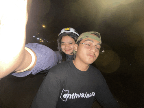 a man wearing a black shirt that says enthusiasm stands next to a woman wearing a helmet