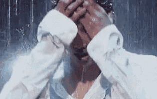a man in a white shirt is standing in the rain covering his face with his hands .