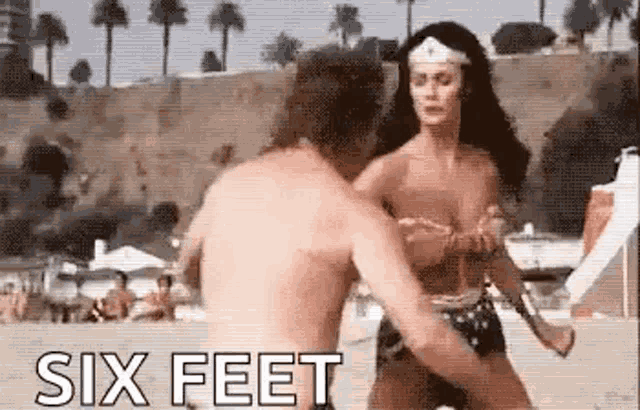 wonder woman is standing next to a shirtless man on a beach .