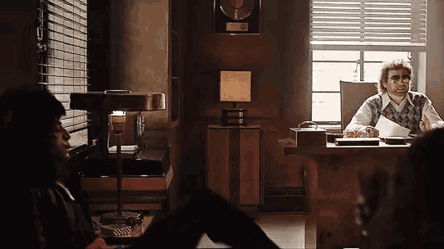 a man is sitting at a desk in a dark room with blinds on the windows .