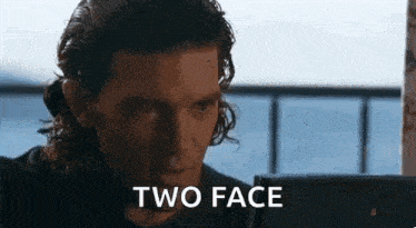 a man with long hair is sitting in front of a laptop computer and says `` two face '' .