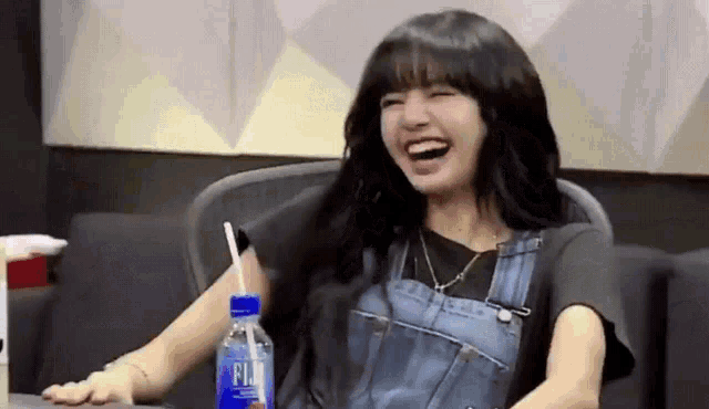 a woman in overalls is laughing while sitting in a chair with a bottle of fiji water .