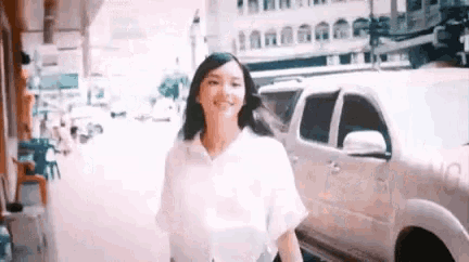 a woman in a white shirt is walking down the street next to a white car .