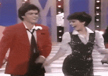 a man in a red jacket and tie is holding a woman 's hand on a stage .