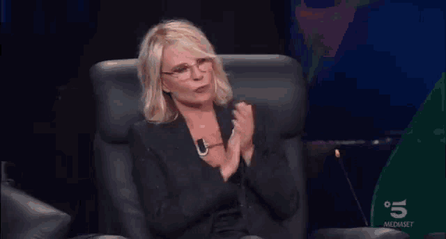 a woman wearing glasses and a black jacket is sitting in a chair and clapping her hands .