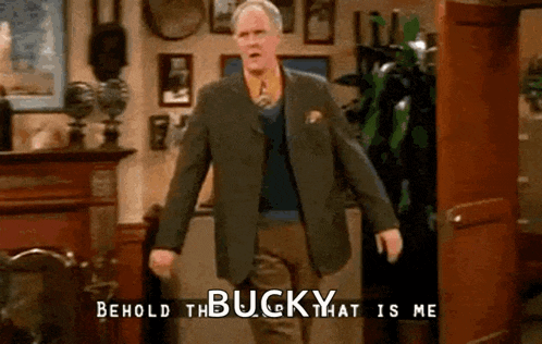 a man in a suit and tie is standing in a living room and says behold the bucky that is me .