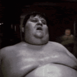 a very fat man without a shirt is sitting in a dark room with his mouth open .