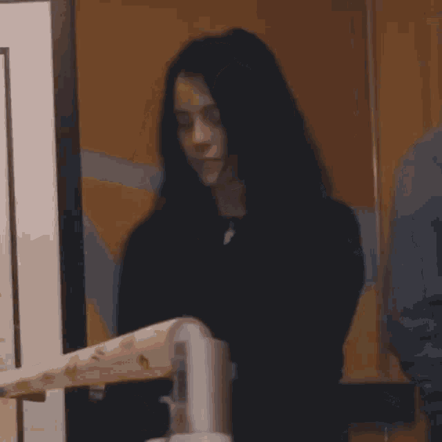 a woman in a black sweater is standing in front of a wall holding a piece of paper .