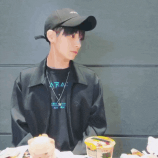 a man wearing a baseball cap and a black jacket is sitting at a table with a cup of food .