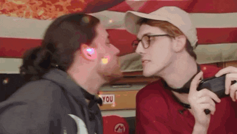 two men are kissing each other in front of an american flag . one of the men is wearing glasses .