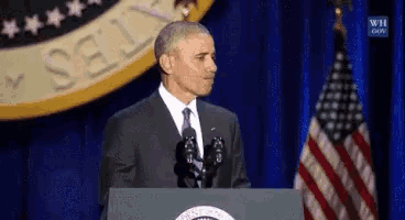 a man in a suit and tie stands at a podium with an american flag in the background