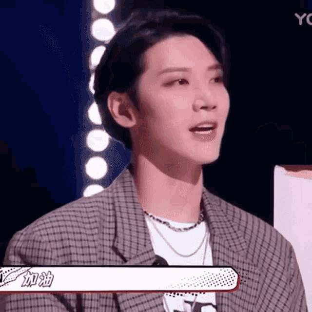 a young man wearing a plaid jacket and a necklace is sitting in front of a microphone .