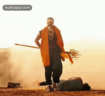 a man in an orange robe is standing next to a man laying on the ground holding a spear .