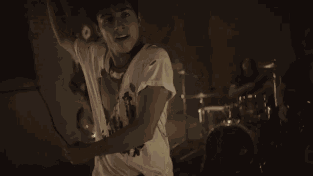 a man in a white t-shirt is standing in front of a drum set and a guitar player
