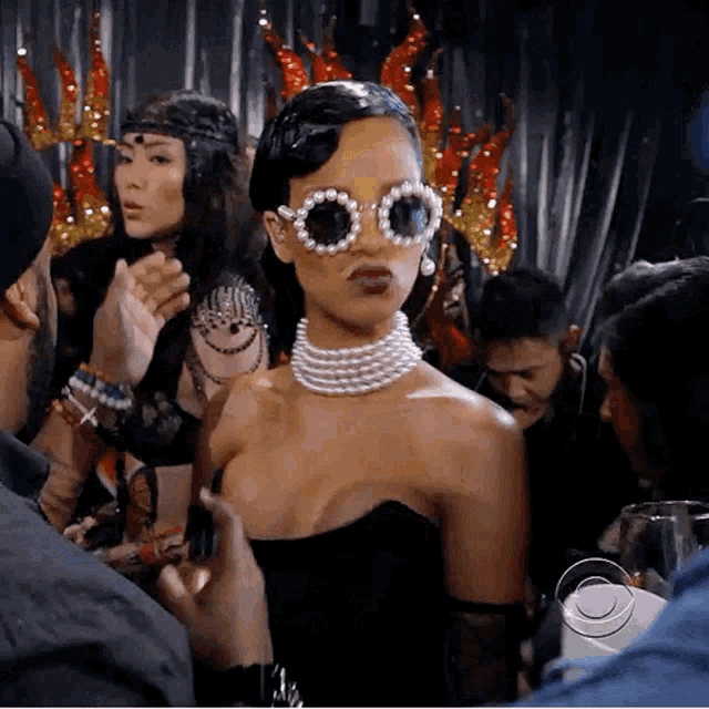 a woman wearing sunglasses and a pearl necklace is surrounded by people