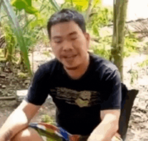 a man in a black shirt is sitting on the ground in the woods .