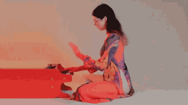 a woman in a colorful dress is kneeling down playing a red piano .