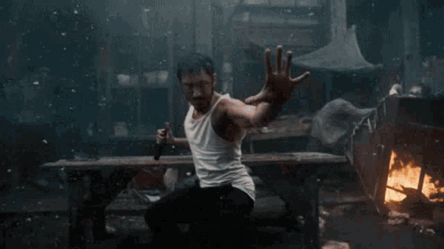 a man in a white tank top is holding a stick in his hand while standing in front of a fire .