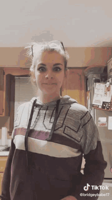 a woman wearing a grey and purple hoodie is standing in a kitchen with her hands on her hips