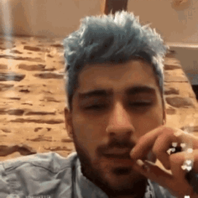 a man with blue hair is laying on a bed smoking a cigarette .