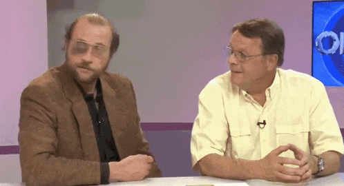 two men are sitting at a table with one wearing glasses and a microphone