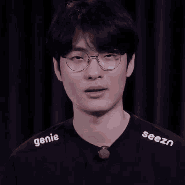 a young man wearing glasses and a black shirt with the word seezn on it is making a fist .