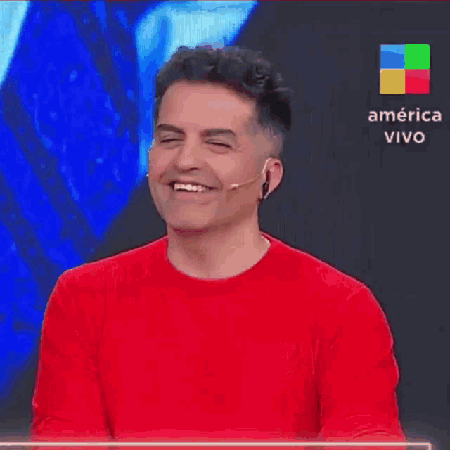 a man in a red sweater is laughing in front of a sign that says america vivo