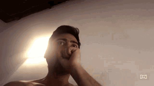 a shirtless man is covering his mouth with his hand in front of a white wall .