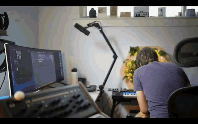 a man in a purple shirt sits at a desk in front of a computer monitor and keyboard
