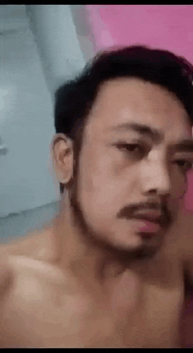 a shirtless man with a beard is taking a selfie in front of a pink wall .