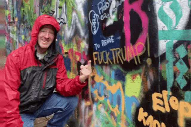 a man in a red jacket giving a thumbs up in front of a wall with graffiti on it that says fuck trump