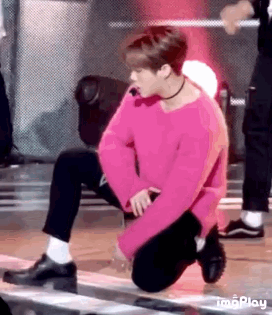 a man in a pink sweater and black pants is kneeling on a stage .