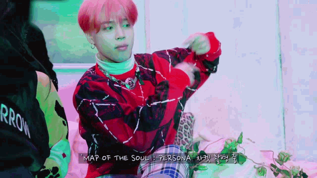 a young man with pink hair is sitting on a couch wearing a red sweater and plaid pants