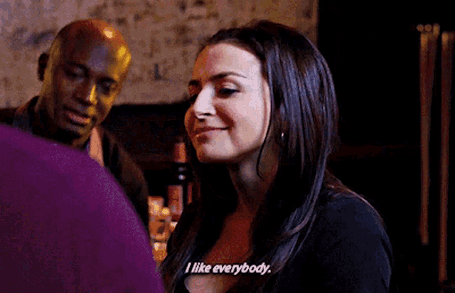 a woman is smiling and saying `` i like everybody '' while talking to a man in a bar .