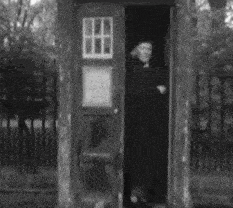 a black and white photo of a man in a black coat standing in a phone booth