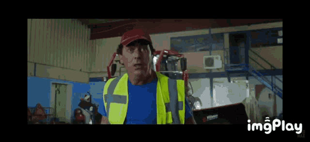 a man wearing a yellow vest and a red hat is standing in front of a computer screen that says imgplay on it