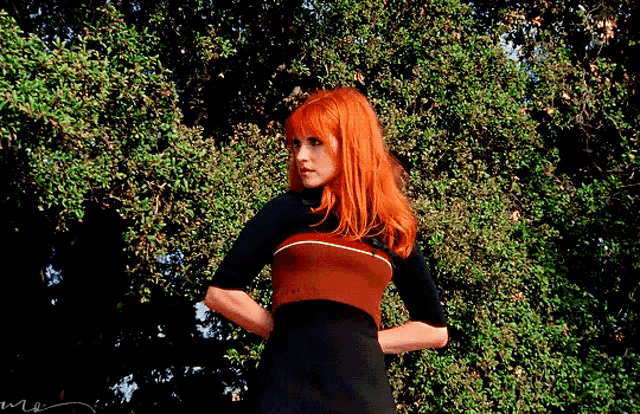 a woman with red hair is standing in front of trees with her hands on her hips