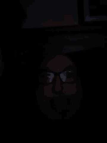 a person wearing glasses in the dark with a picture in the background