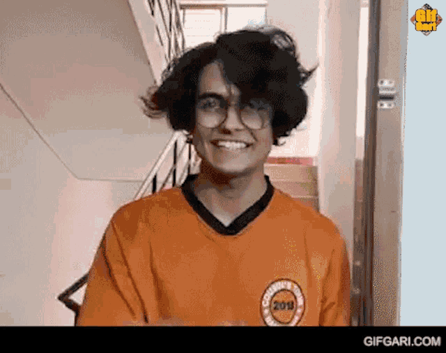 a young man wearing glasses and an orange shirt is smiling and looking at the camera .
