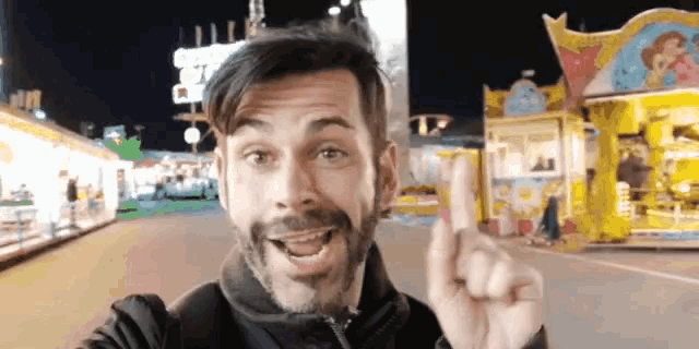 a man with a beard is smiling and pointing up in front of a carnival ride