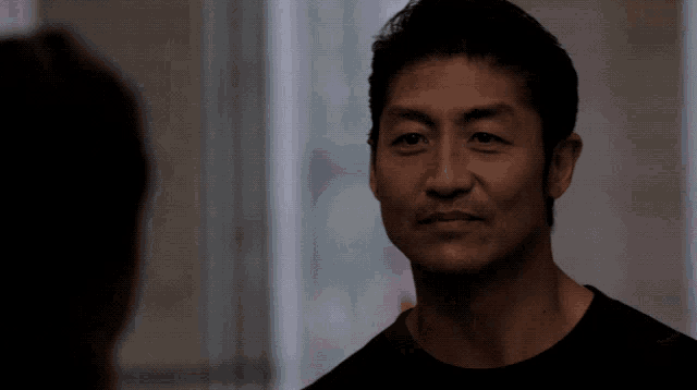 a man in a black shirt looks at another man in a dark room