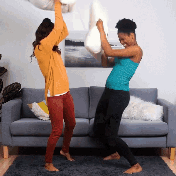 two women pillow fight in front of a couch