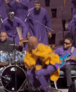 a man in a yellow jacket is sitting on a stool on a stage playing drums .