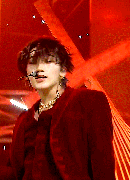 a man wearing a red jacket and a gold necklace is singing into a microphone on stage .