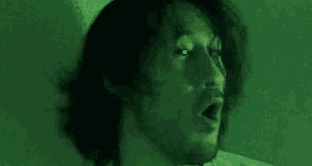 a man with long hair and a beard is making a funny face with his mouth open in a green light .