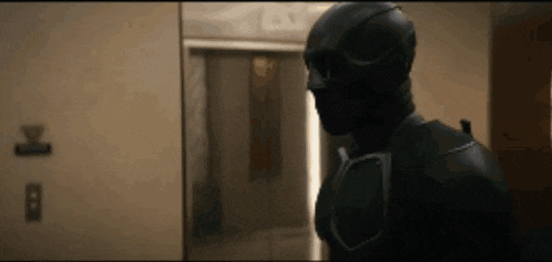 a man in a black superhero costume is standing in a hallway .