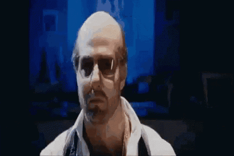 a bald man wearing sunglasses and a white shirt is standing in front of a blue wall .